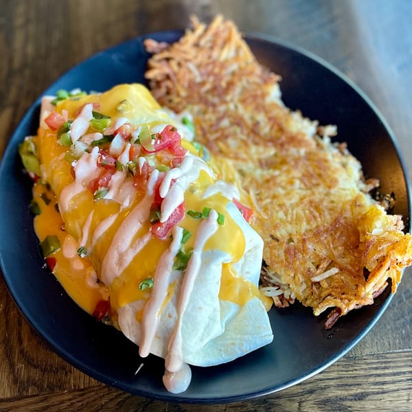 A breakfast burrito topped with cheese, salsa, and sauce, served with crispy hash browns on a dark plate.