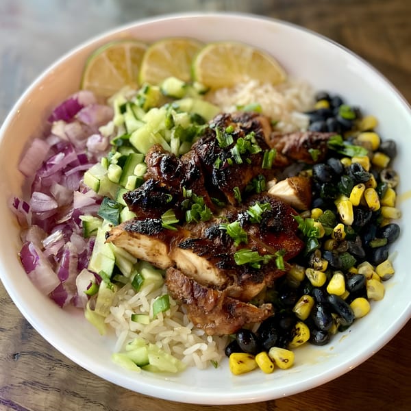 A vibrant chicken bowl with rice, black beans, corn, red onion, cucumber, and lime wedges.